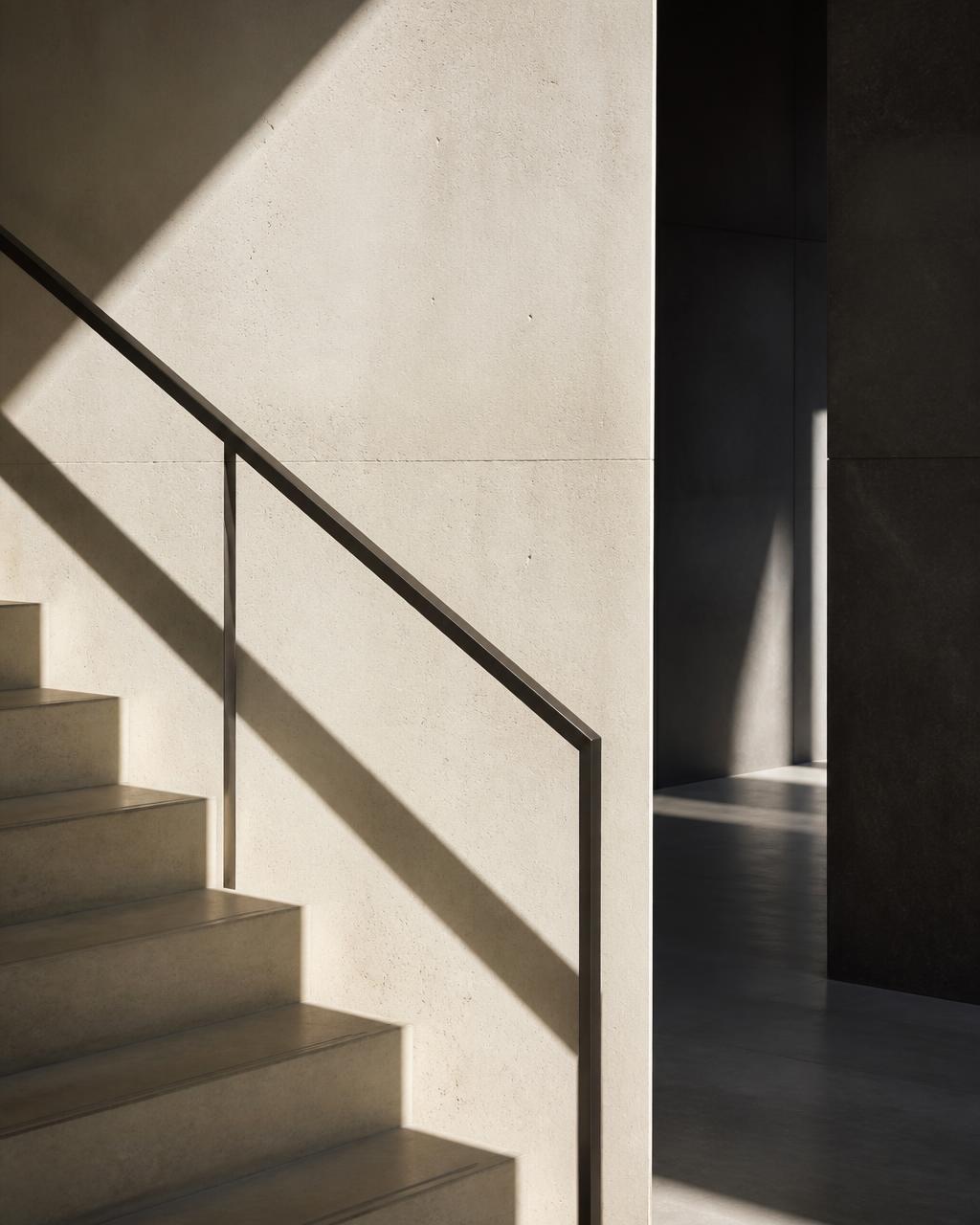 Concrete stair edge and slim bronze handrail casting a diagonal shadow in a quiet commercial lobby.